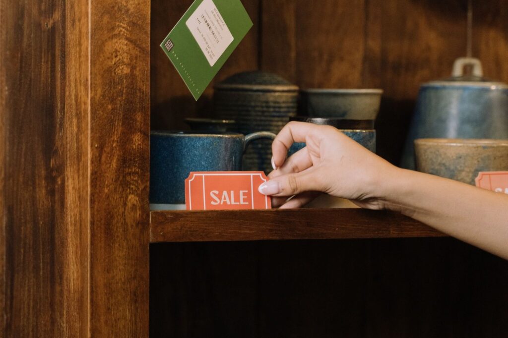 A hand setting a red sale tag on a wooden shop shelf beside pottery