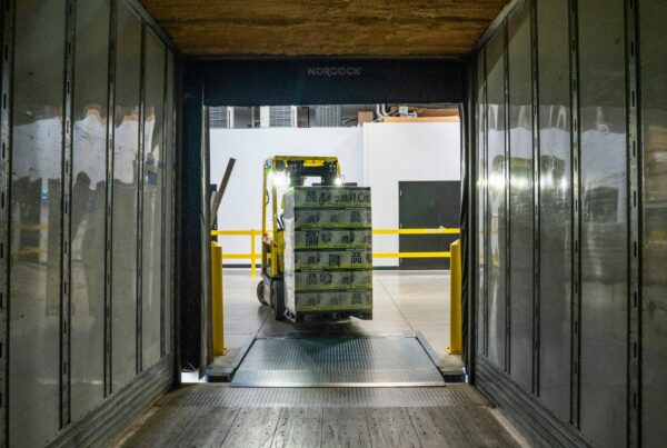 A forklift carrying a pallet of boxes toward a trailer at a warehouse loading dock