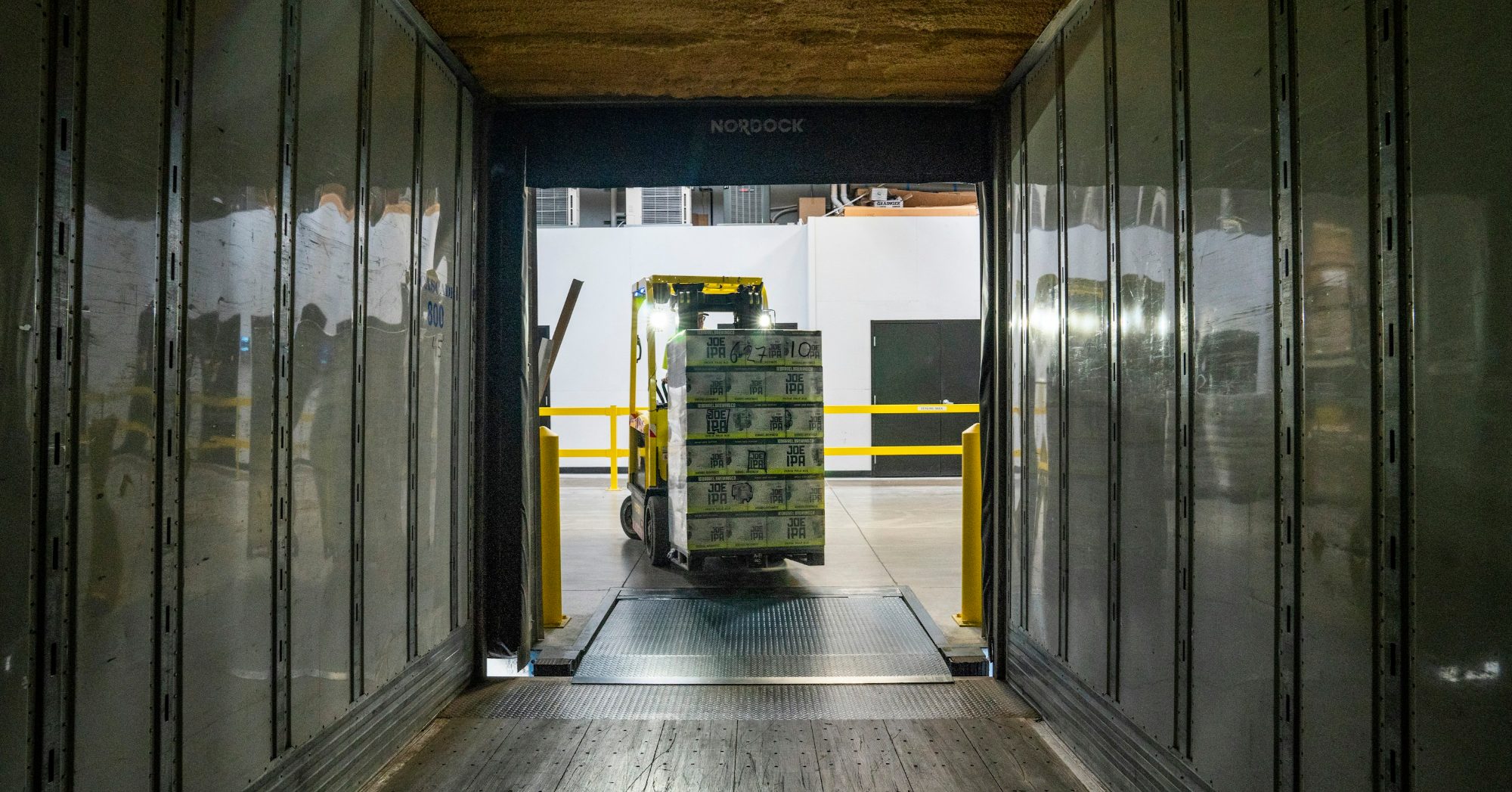 A forklift carrying a pallet of boxes toward a trailer at a warehouse loading dock