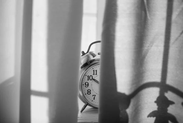 Black and white photo of a vintage alarm clock standing behind a sheer curtain in low light