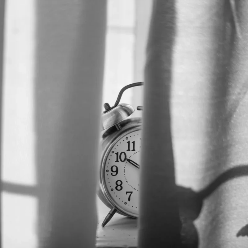 Black and white photo of a vintage alarm clock standing behind a sheer curtain in low light