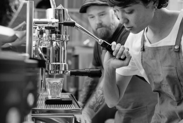 A young barista working the espresso machine while a senior colleague watches from behind the counter