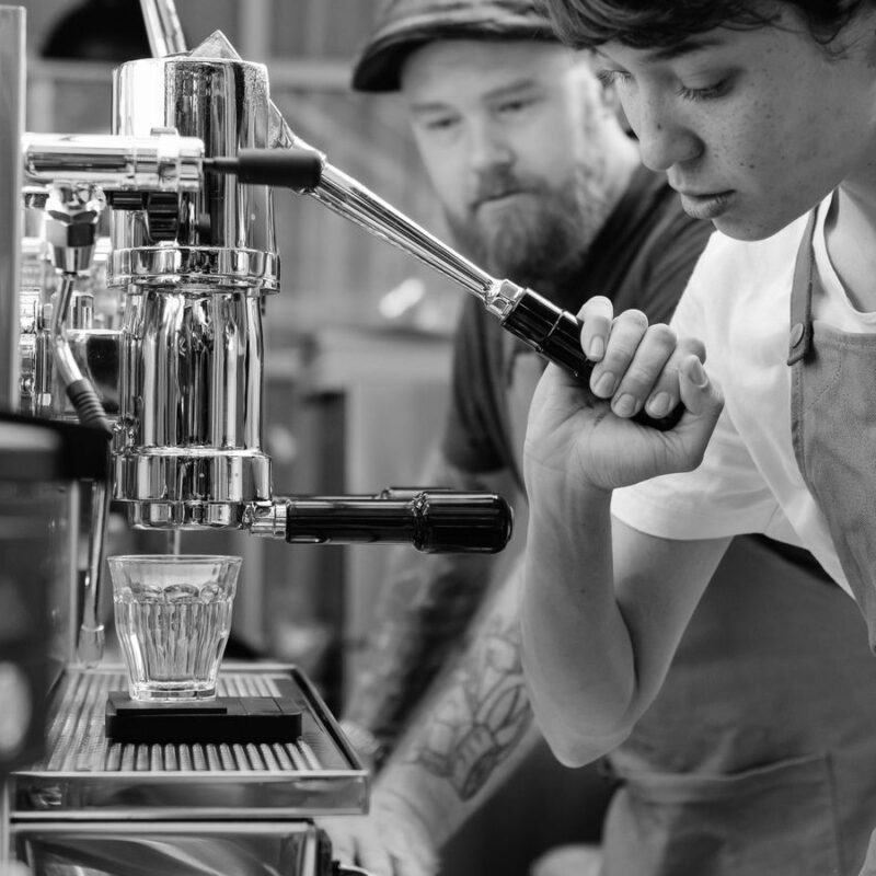 A young barista working the espresso machine while a senior colleague watches from behind the counter