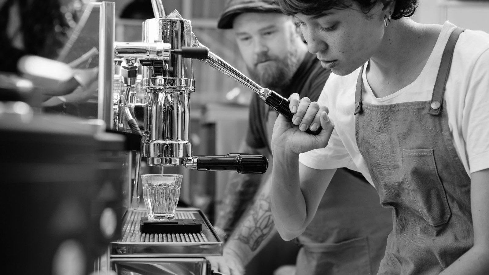 A young barista working the espresso machine while a senior colleague watches from behind the counter
