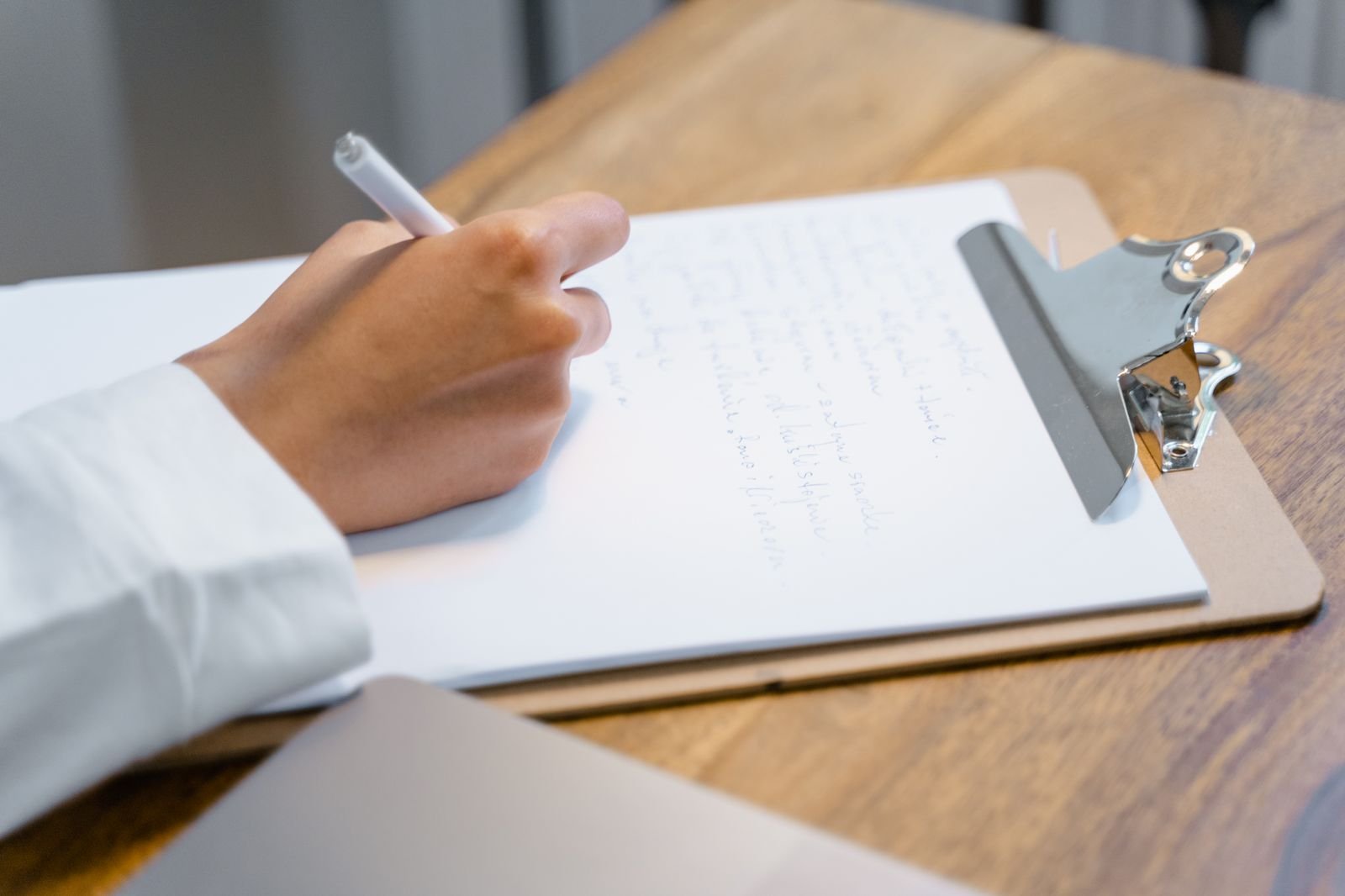 A hand writing entries on a sheet clipped to a clipboard on a wooden table