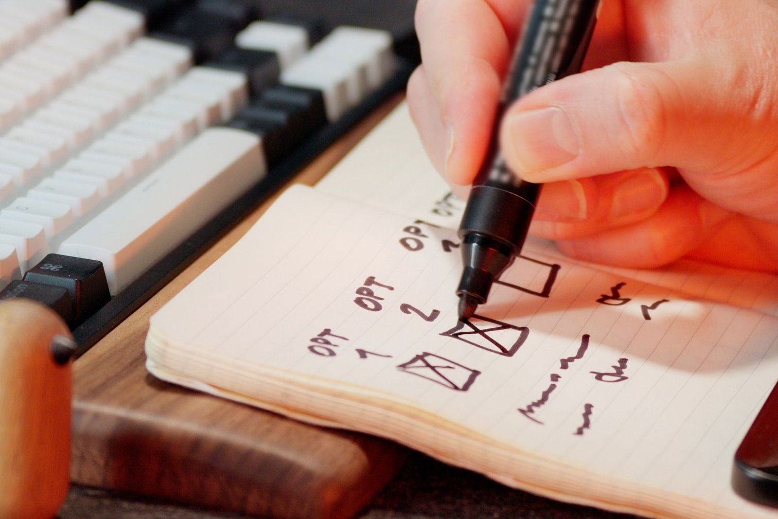 A hand crossing items off a handwritten list in a notebook beside a keyboard