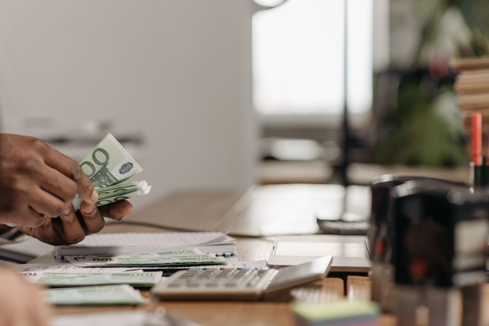 Hands counting paper money at an office desk beside a calculator.