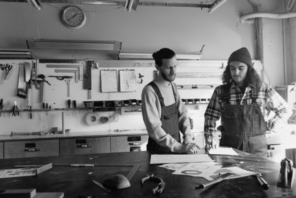 Two craftsmen at a workshop table, looking over plan sheets spread in front of them