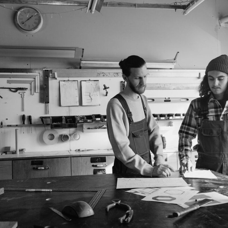 Two craftsmen at a workshop table, looking over plan sheets spread in front of them