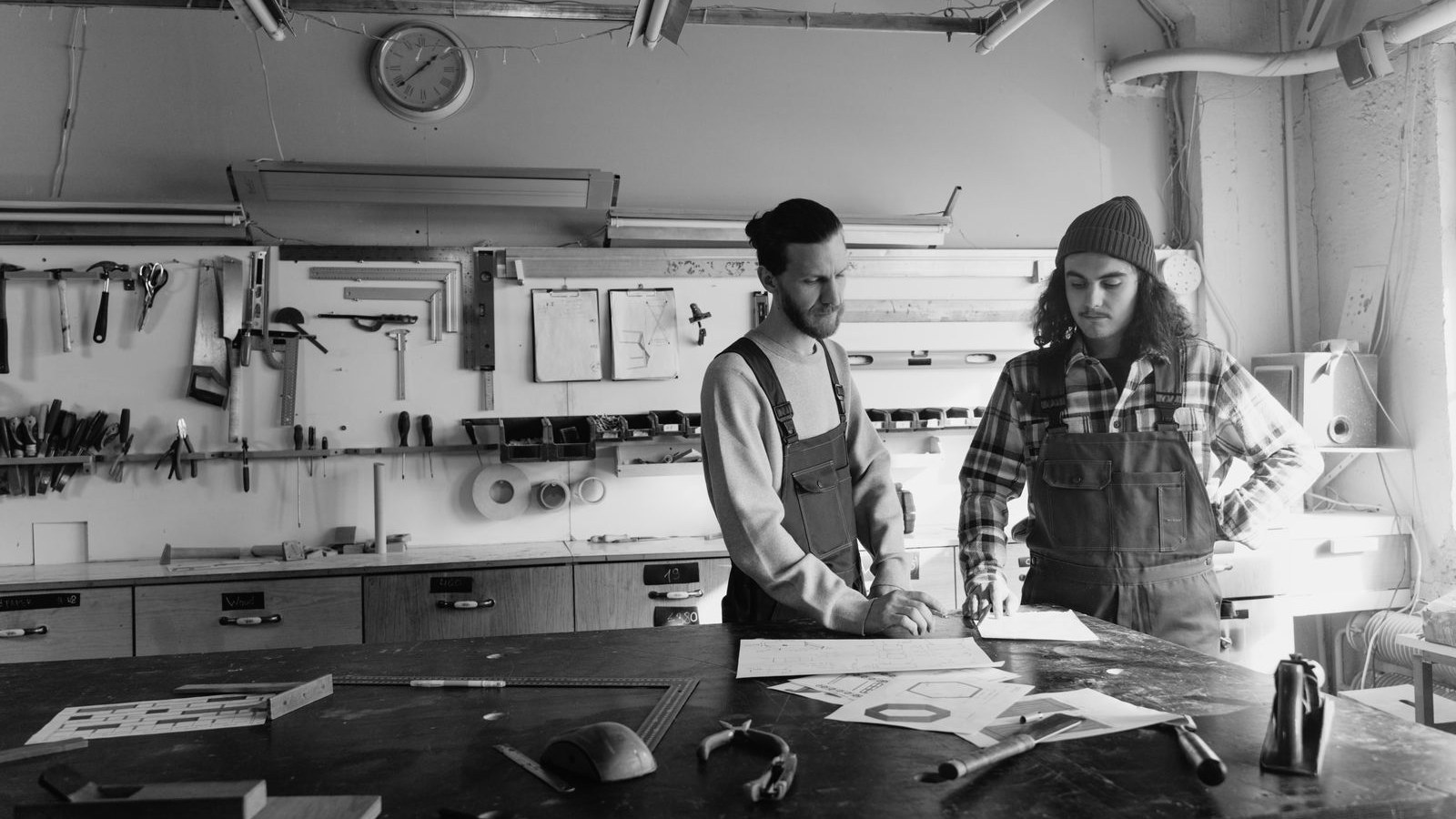 Two craftsmen at a workshop table, looking over plan sheets spread in front of them