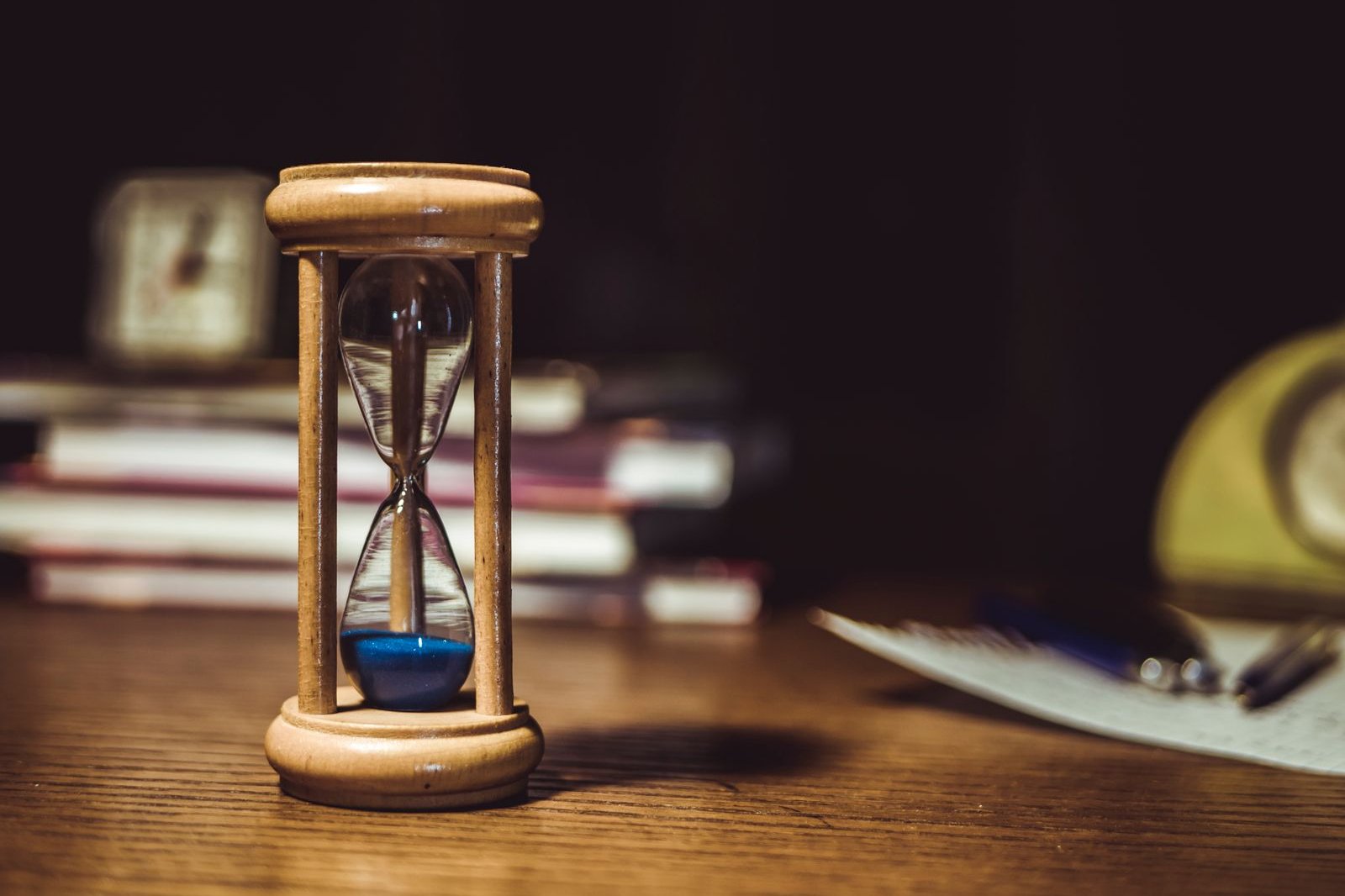 A wooden hourglass with blue sand on a desk, with books behind it