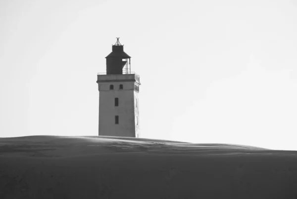A lighthouse standing alone on a dark sand dune against the sky, in black and white