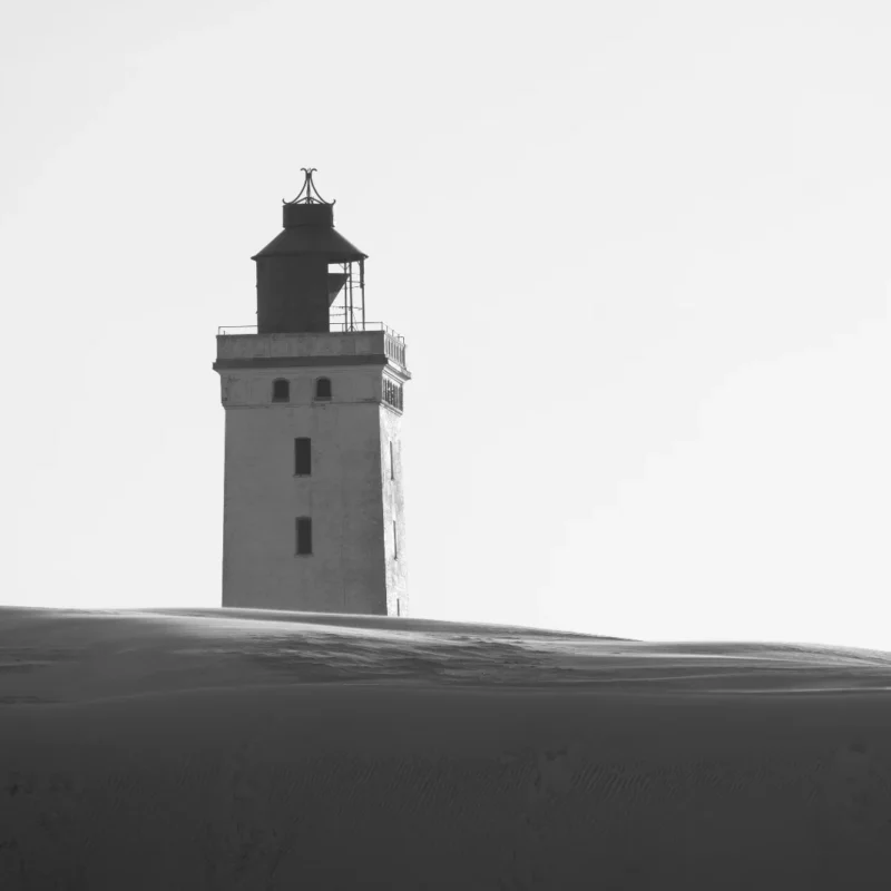 A lighthouse standing alone on a dark sand dune against the sky, in black and white