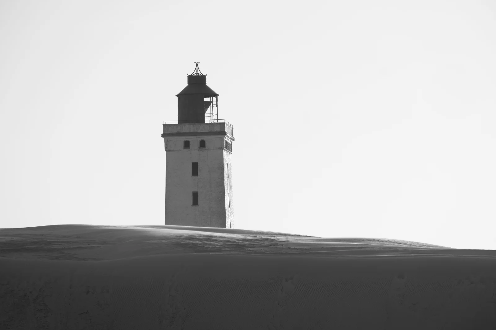 A lighthouse standing alone on a dark sand dune against the sky, in black and white