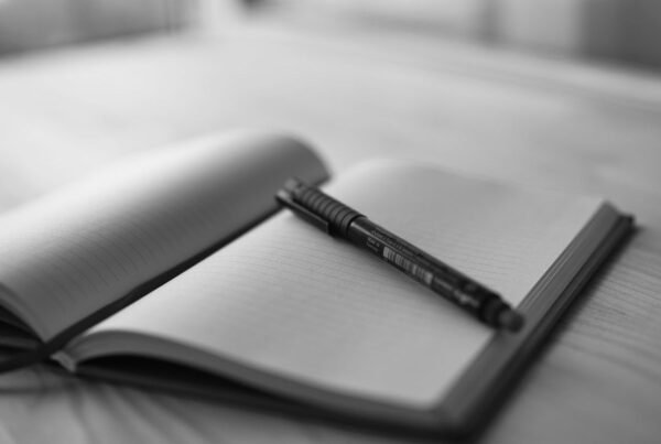 An open bound notebook with ruled pages and a pen resting on it, on a wooden desk