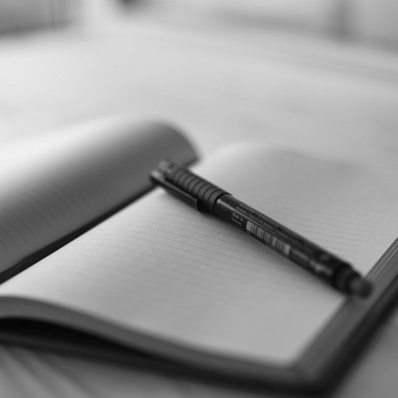 An open bound notebook with ruled pages and a pen resting on it, on a wooden desk