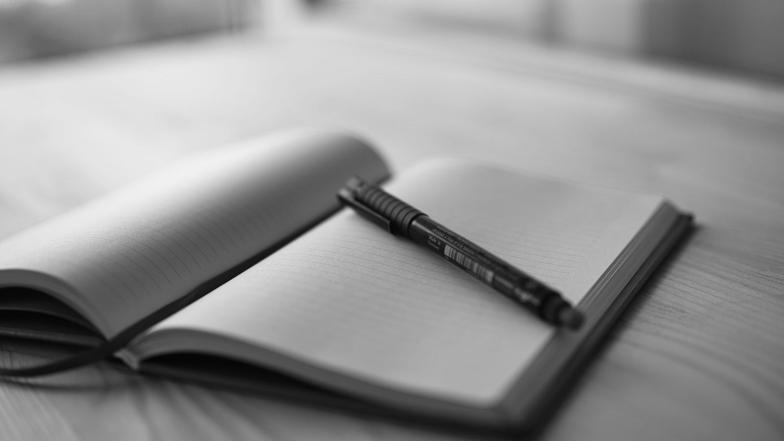 An open bound notebook with ruled pages and a pen resting on it, on a wooden desk