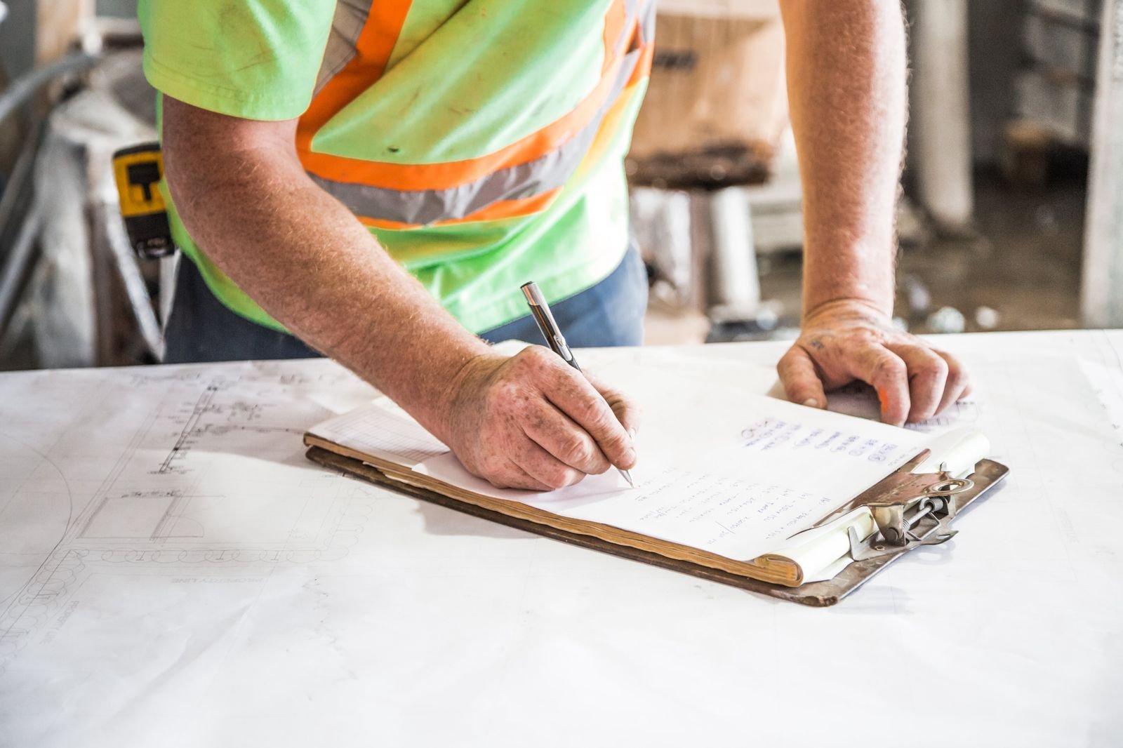 A worker writing by hand on a clipboard, a large plan spread on the table under it